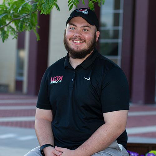 A man with a beard wearing a black "网赌最好最大平台 Soccer" shirt and cap is sitting outdoors, smiling at the camera.